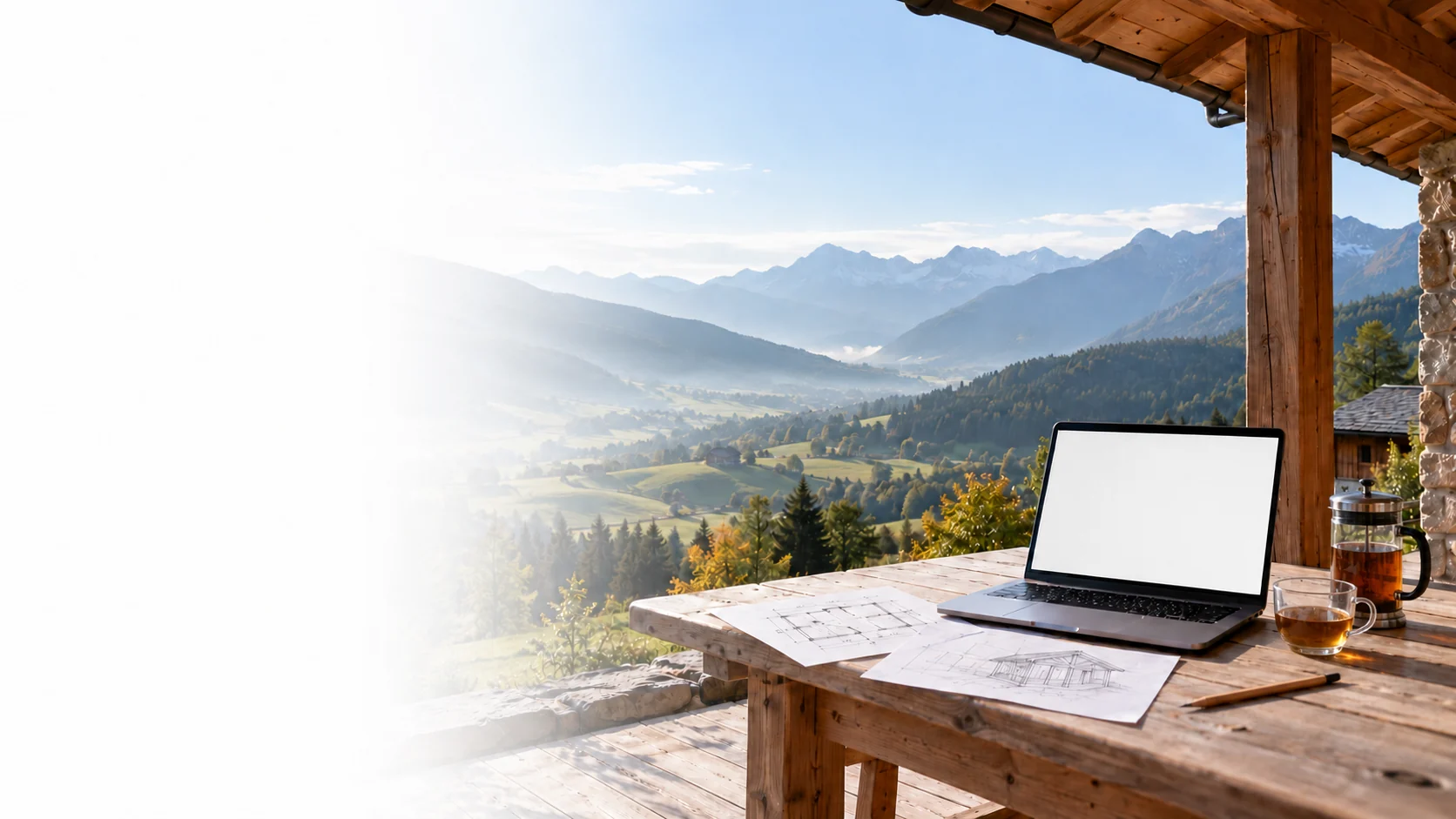 A laptop, hand-drawn plans and glass tea cups on a wooden terrace overlooking a mountain valley.