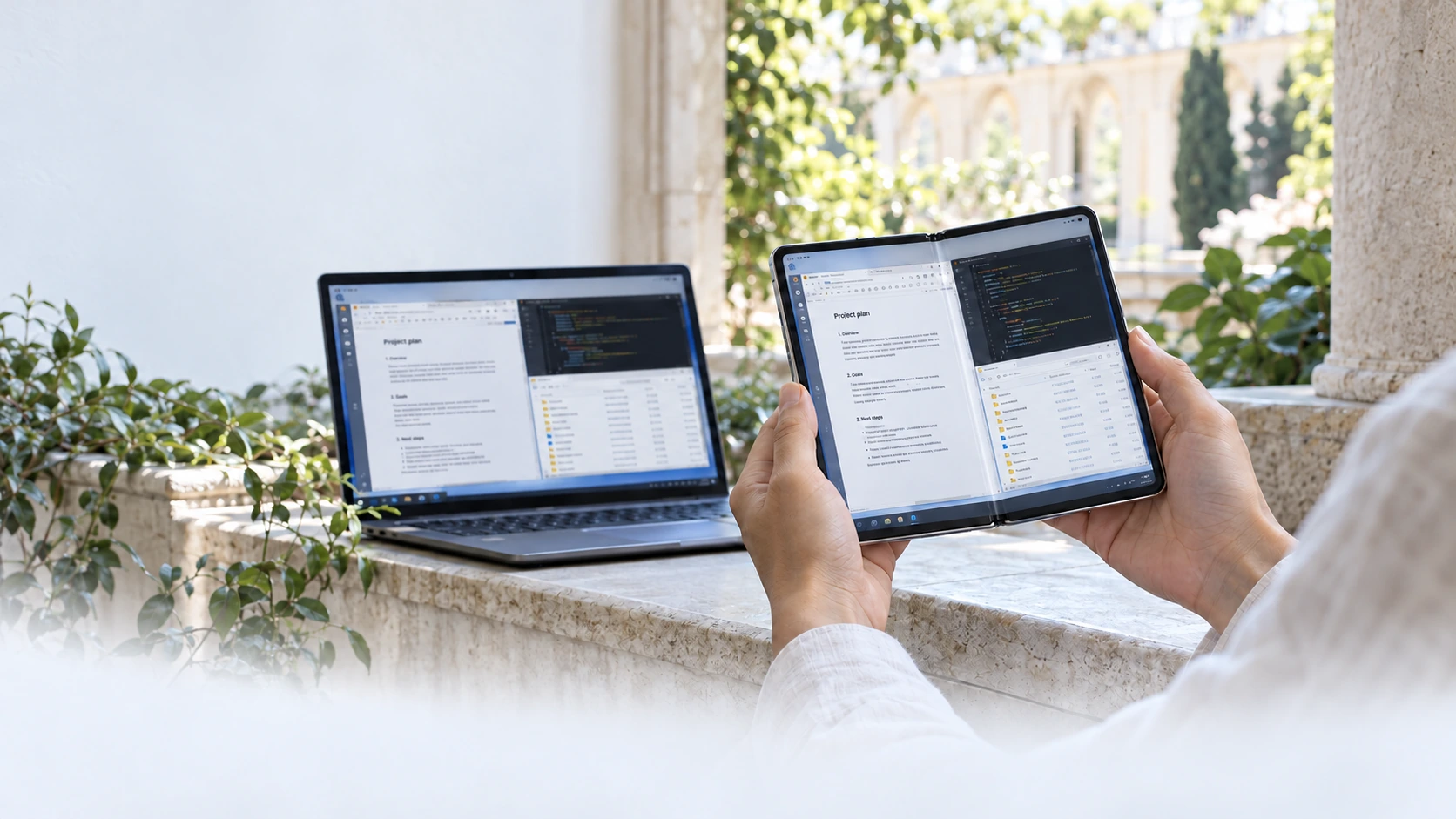 Hands hold an unfolded book-style device in a sunlit garden arcade. It and the laptop on the ledge show the same document, code editor and files: an illustration of different ways to access a workspace.