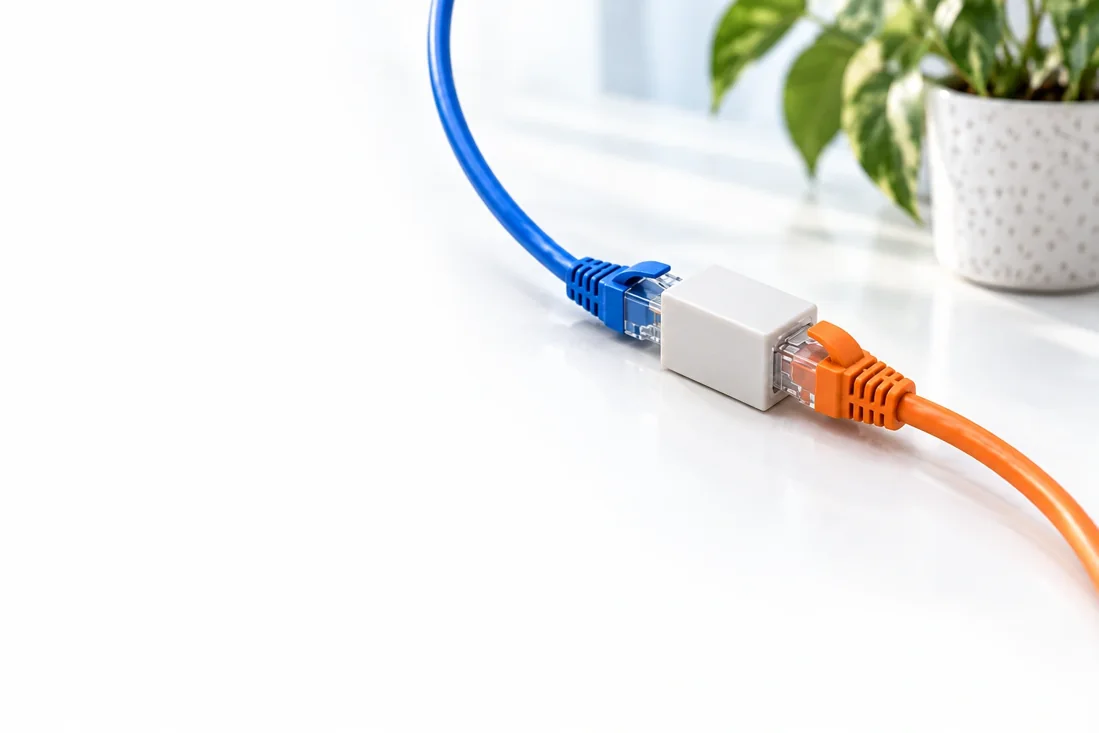 A blue and an orange network cable connected through one white inline coupler on a sunlit desk.